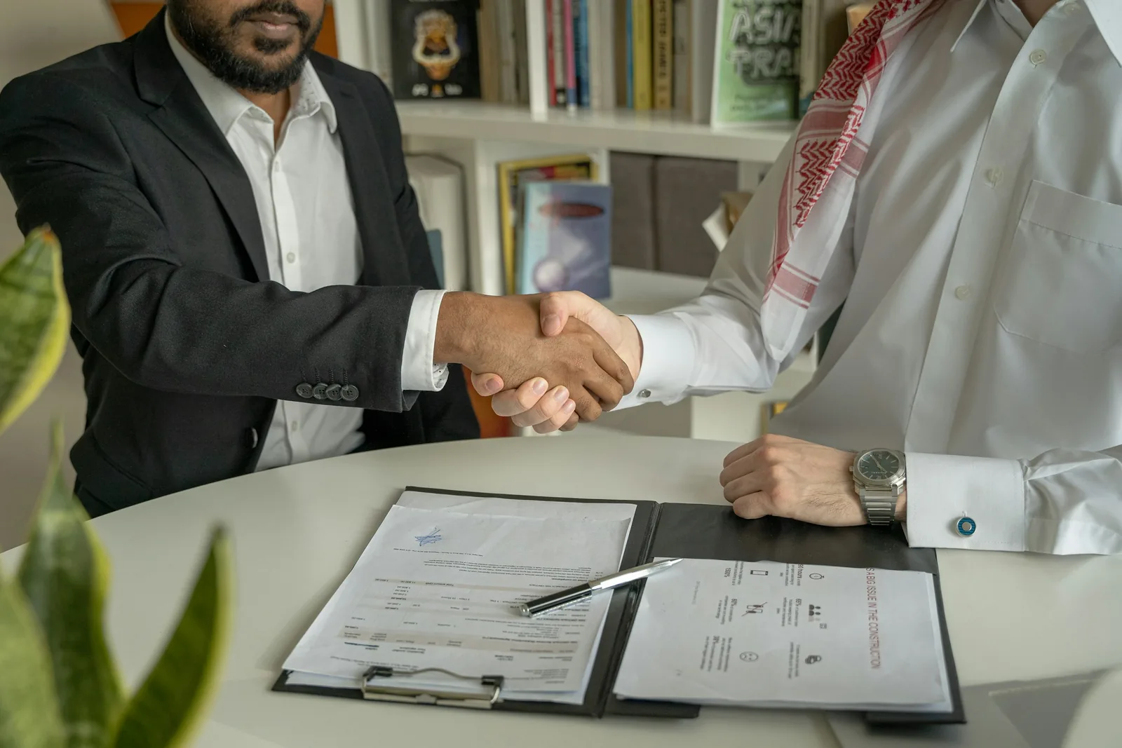 Two men shaking hands across an office desk with papers on it
